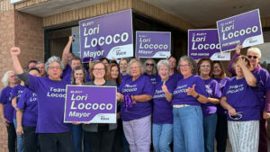 People in purple shirts holding up elect lori lococo signs in front of building